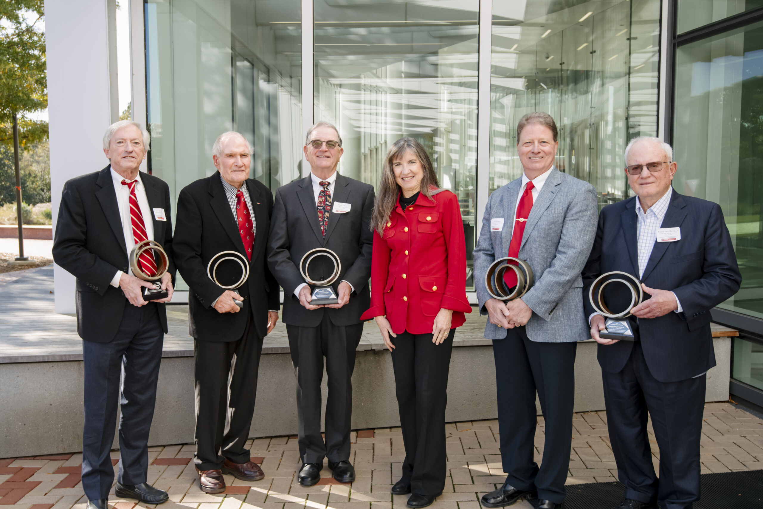 A full-body photo of six people—five men and one woman—standing side-by-side outdoors in front of a modern building with large glass windows. The five men are all dressed in dark suits, and each is holding a trophy shaped like a stylized ring on a base. The woman, standing slightly to the right of the center, is wearing a bright red blazer and black pants. They appear to be posing for a photograph, possibly at an awards or hall of fame event, as indicated by the small identification badges on their lapels.