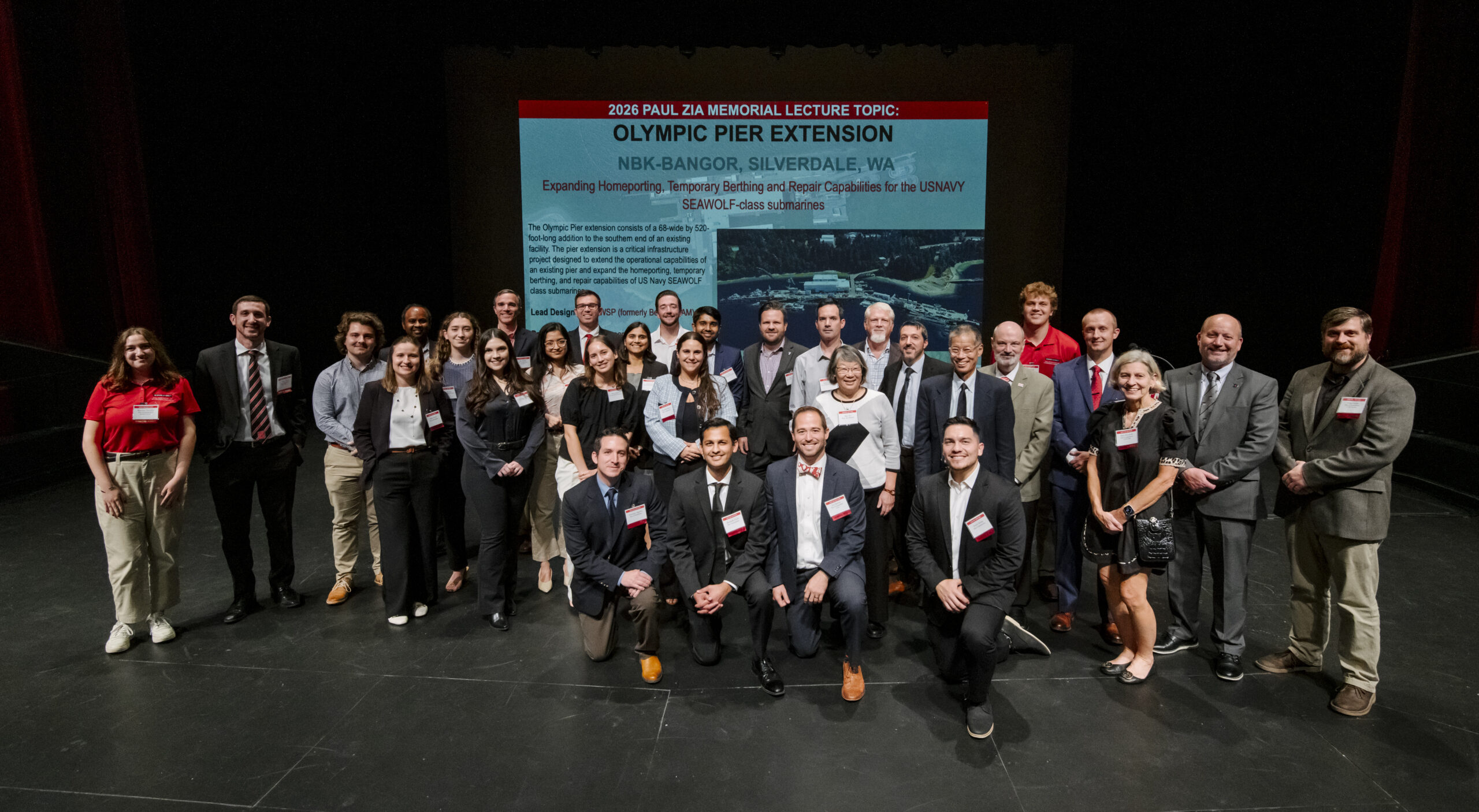 A large group of approximately 30 people, appearing to be professionals and students, stand and kneel on a stage, posing for a group photograph. The people are dressed in business attire or business casual, with some wearing event lanyards. Behind them is a large projection screen displaying text for the "2026 PAUL ZIA MEMORIAL LECTURE TOPIC: OLYMPIC PIER EXTENSION" for "NBK-BANGOR, SILVERDALE, WA". The project involves "Expanding Homeporting, Turnover, Berthing and Repair Capabilities for the US Navy SEA WOLF-Class Submarines." The text also includes a brief project description and credits the "Lead Designer: WSP USA" and "Lead Builder: Kiewit-General." A black and white photo of the pier is visible behind the text on the screen. The stage is dark, focusing the light on the people and the screen.
