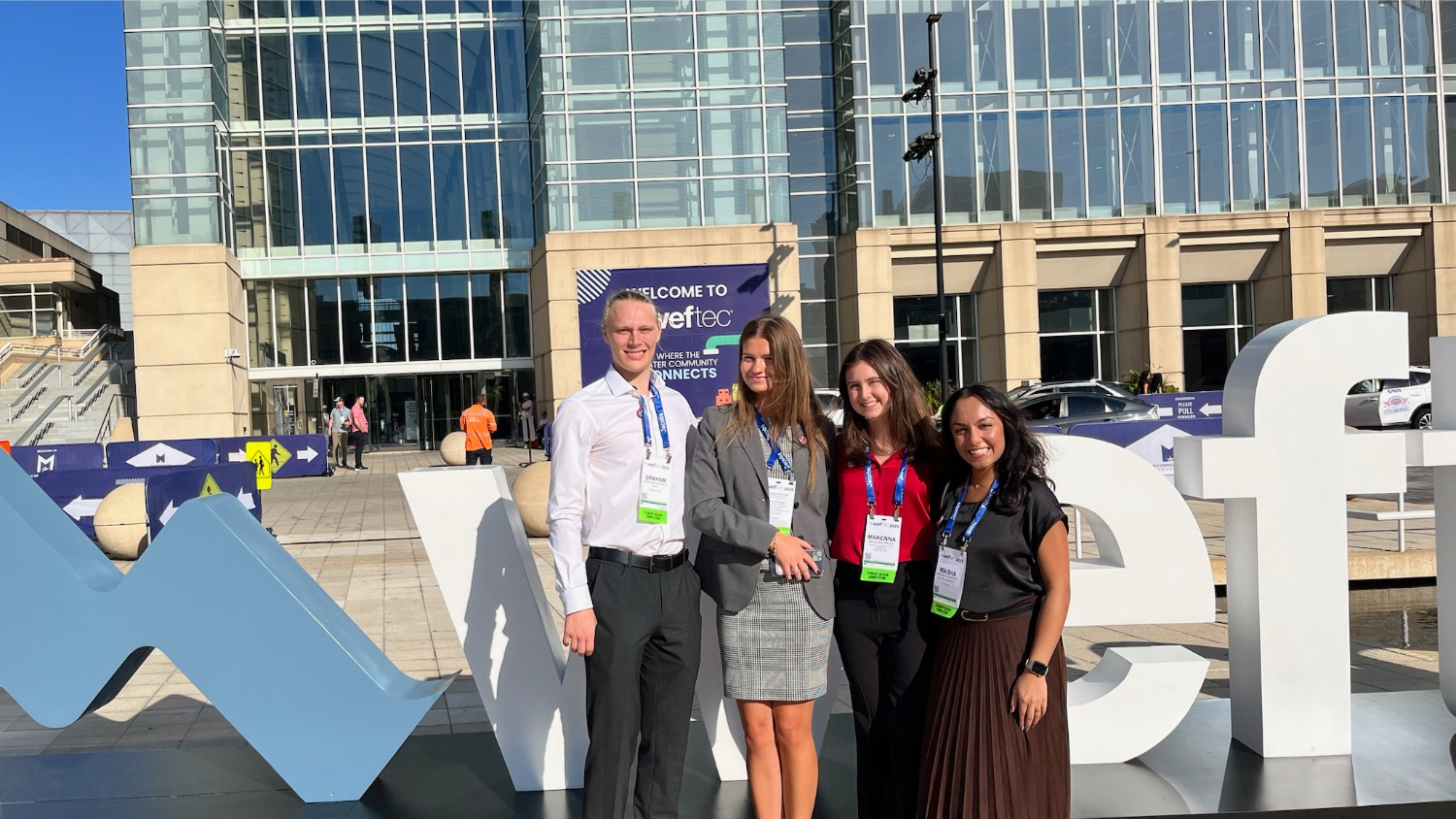 A group of four young professionals (one man and three women) stand smiling in front of a large, modern glass building, posing next to large, white sculptural letters that spell "WEF" with a stylized blue 'W' and a smaller sign visible behind them that says "WELCOME TO WEFTEC."