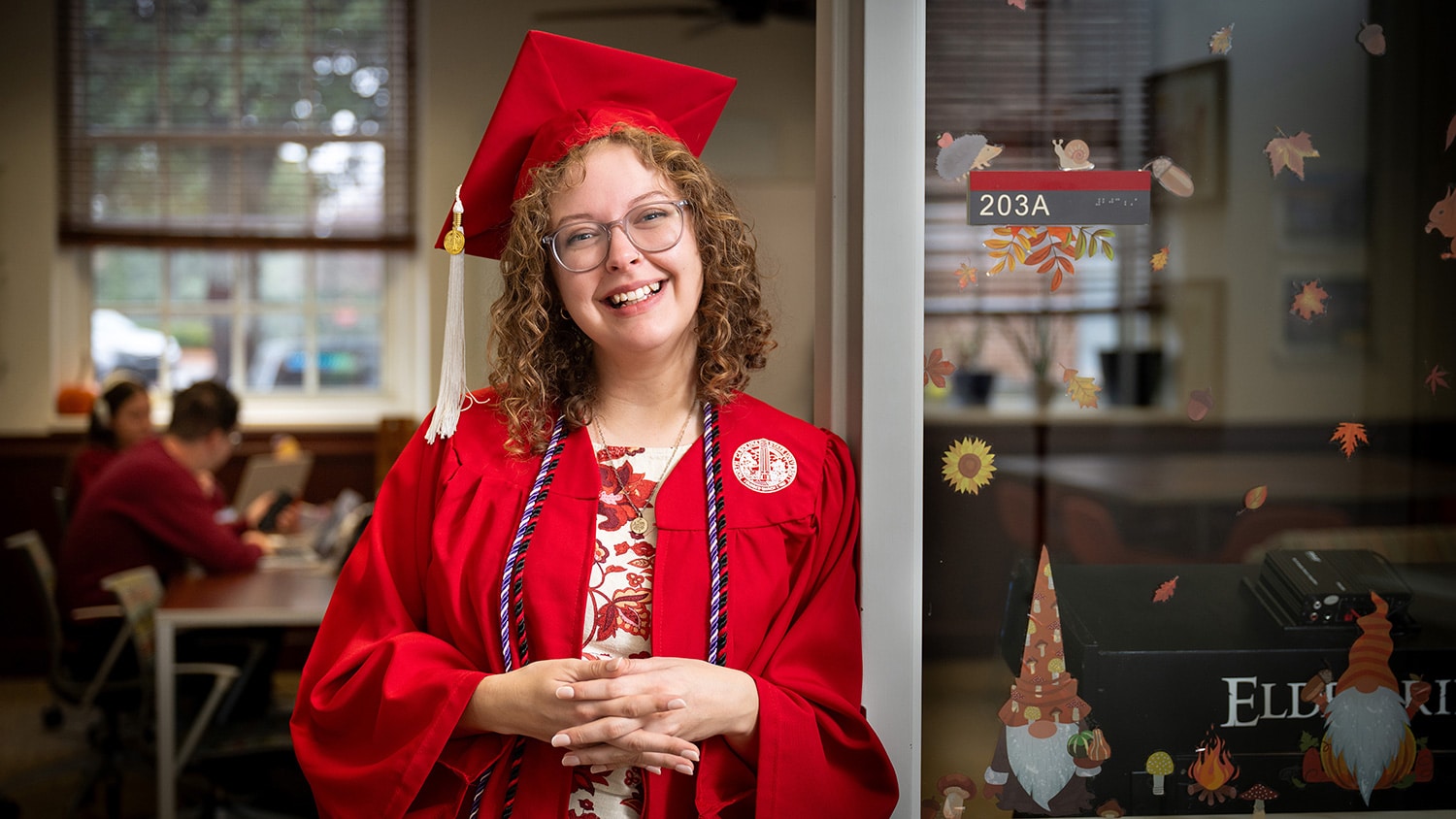 A medium shot of a smiling young woman with curly blonde hair and glasses, wearing a bright red graduation gown and mortarboard cap with a white tassel. She is leaning against a white doorframe in an indoor setting.