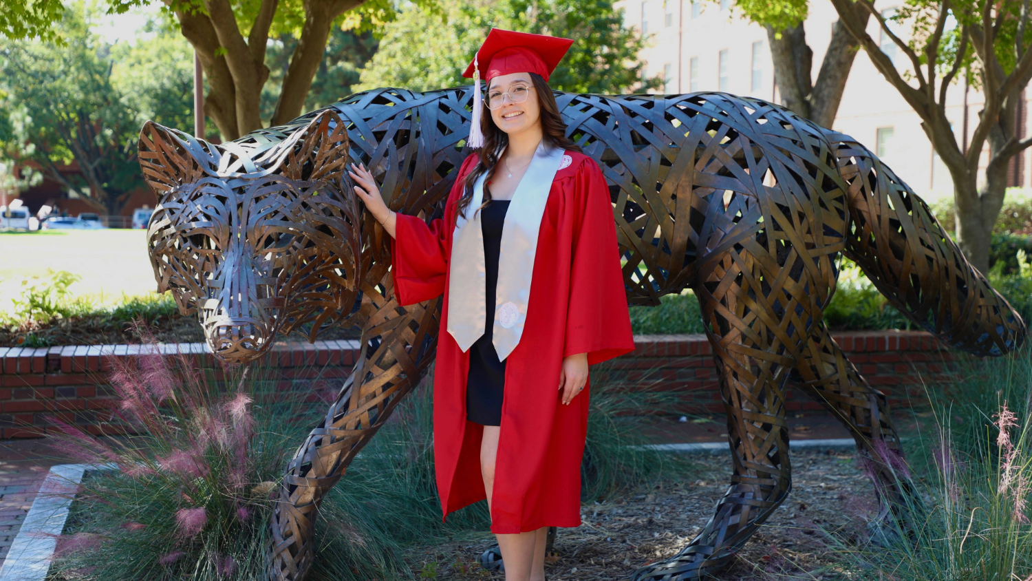 A young woman with glasses, wearing a bright red graduation cap and gown with a white stole, is standing outdoors next to a large, metallic, sculpted wolf statue. She is smiling and gently touching the statue with her right hand. The background features green trees, some brickwork, and pinkish-purple ornamental grass.