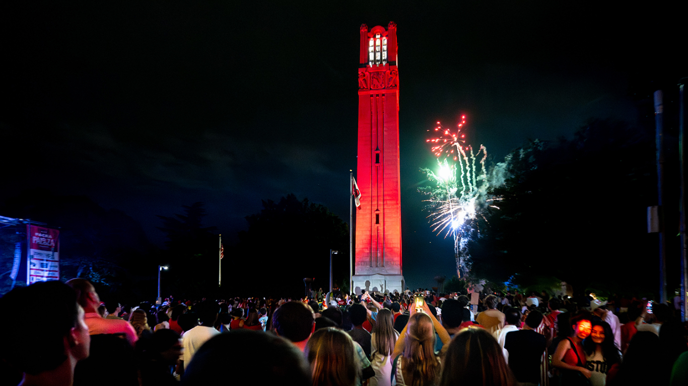 A photo of NC State’s belltower, lit up in red with fireworks in the background.